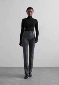 Fekete garbó felső szürke, testre szabott nadrággal párosítva, amely slim fit, magas derekú és fényes fekete bokacsizmát visel. Egyszerű háttér.