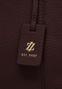 Etichetta in pelle marrone scuro con texture, logo in oro impresso e "EST. 2007" attaccata a una superficie in pelle coordinata con cuciture visibili.