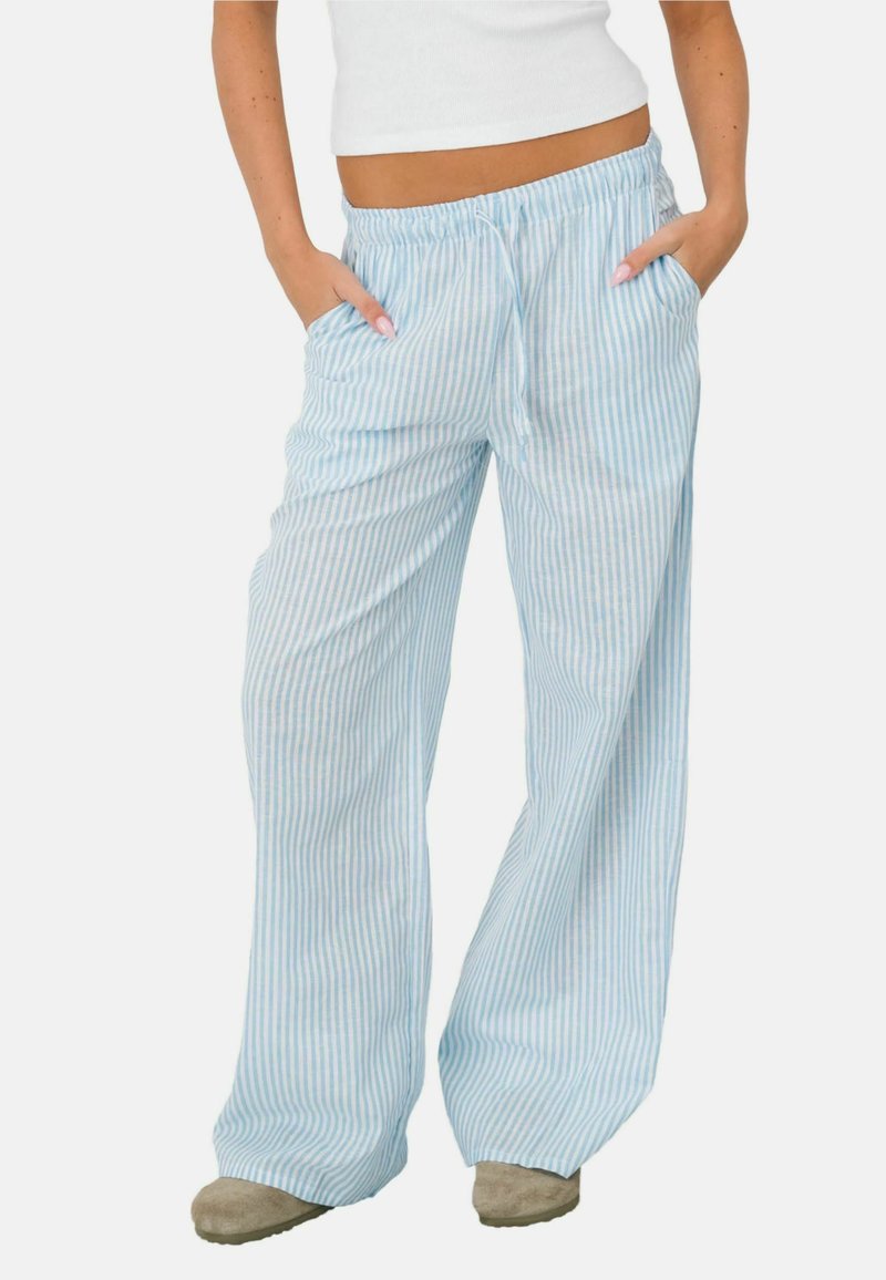 Femme portant un pantalon ample à rayures verticales bleu clair et blanches, avec une ceinture élastique et des poches, associé à des chaussures beiges et un haut blanc.