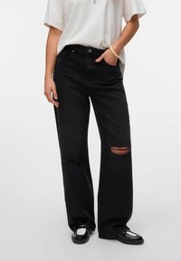 Svarta vida jeans med slitna områden och synliga hål. Materialet är denim, i kombination med en lös vit T-shirt och svarta skor.