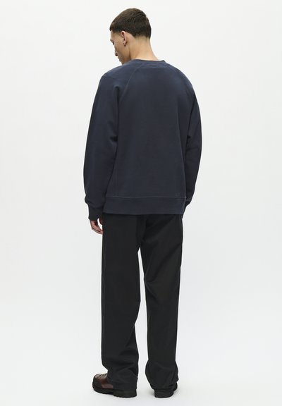 Moški stoji z hrbtom proti gledalcu, oblečen v temno modro jopa s kapuco, črne široke hlače in rjave čevlje, na enostavni svetli podlagi.