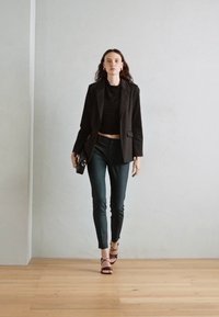 Černý blejzr přes černý top, doplněný tmavě zelenými slim-fit kalhotami a černými sandály na vysokém podpatku. Modelka drží malou černou kabelku.