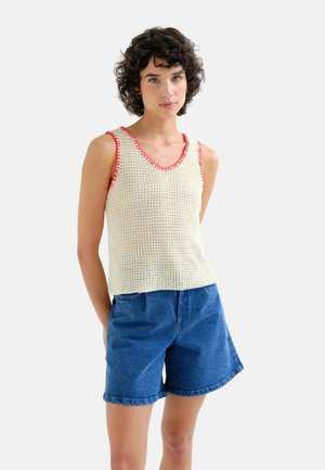Mujer con cabello corto y rizado, que lleva un top de punto sin mangas de color crema con ribete rojo y pantalones cortos vaqueros azules, de pie frente a un fondo liso.