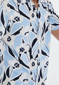 Kurzärmliges Hemd mit Knopfleiste, das einen hellblauen Hintergrund, große weiße Blumen und dunkelblaue Akzente aufweist. Es hat einen lässigen Schnitt und eine glatte Textur.