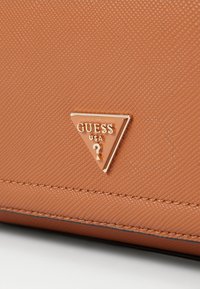 Handbag dengan bahan sintetis tekstur oranye dengan emblem emas segitiga yang menampilkan "GUESS USA" dan tanda tanya. Jahitan yang kokoh terlihat.