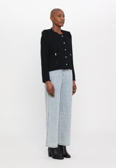 Schwarze gewebte Jacke mit ausgefransten Kanten, silbernen Knöpfen und zwei Reißverschluss-Taschen, kombiniert mit hellblauen geraden Jeans und schwarzen Stiefeletten.