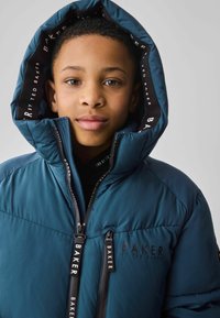 Veste rembourrée en bleu sarcelle, avec un col haut, un logo sur la poitrine et des tirettes de fermeture éclair noires avec une marque. Capuche doublée de tissu imprimé.