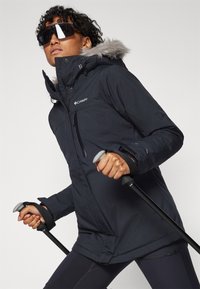 Chaqueta aislante negra con capucha forrada de pelo sintético, bolsillos con cremallera y guantes antideslizantes. Incluye bastones de senderismo, mostrando un diseño sport.