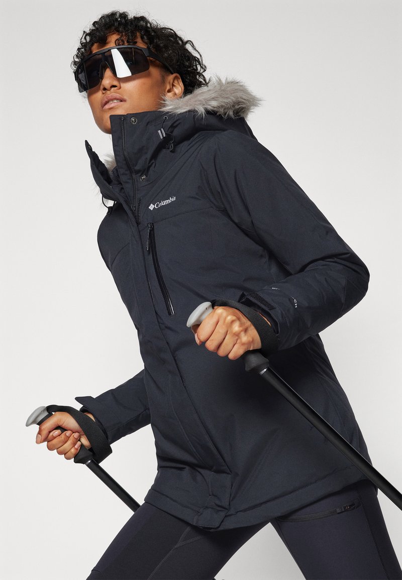 Schwarze isolierte Jacke mit einer Kapuze aus Kunstfell, Reißverschlusstaschen und Grip-Handschuhen. Enthält Trekkingstöcke und zeigt ein sportliches Design.