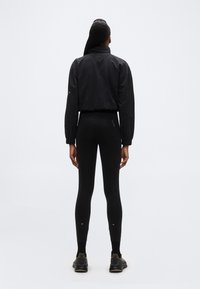 Γυναίκα που στέκεται με τ ην πλάτη, φοράει μαύρο κοντό μπουφάν, μαύρα κολάν, μαύρες κάλτσες και μαύρα αθλητικά παπούτσια με ελαιώδεις αποχρώσεις.