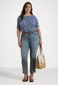Blomstrete blå topp med korte ermer, lyseblå jeans, brun belte, brune kilehæler, holder en flettet veske med et hvitt håndkle oppå.