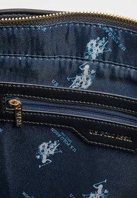 Cartera negra con cremallera, interior de tela con un patrón de logotipos azul y blanco de U.S. Polo Assn., herrajes de cremallera dorados y textura suave.