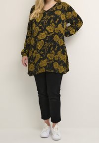 Zwarte bloemen tuniek met mosterdgele rozenpatroon, voorzien van een V-hals en lange mouwen. Gepaard met zwarte broeken en witte sneakers.