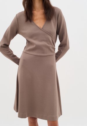 Woman wearing a long-sleeve, taupe-colored wrap dress with a V-neckline, hands resting behind her back against a plain background.