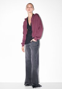 Žena oblečená v purpurovej mikine na zips, čiernom topu s výstrihom do V, širokých vyblednutých čiernych džínsach a čiernych špicatých topánkach, stojaca na bielom pozadí.