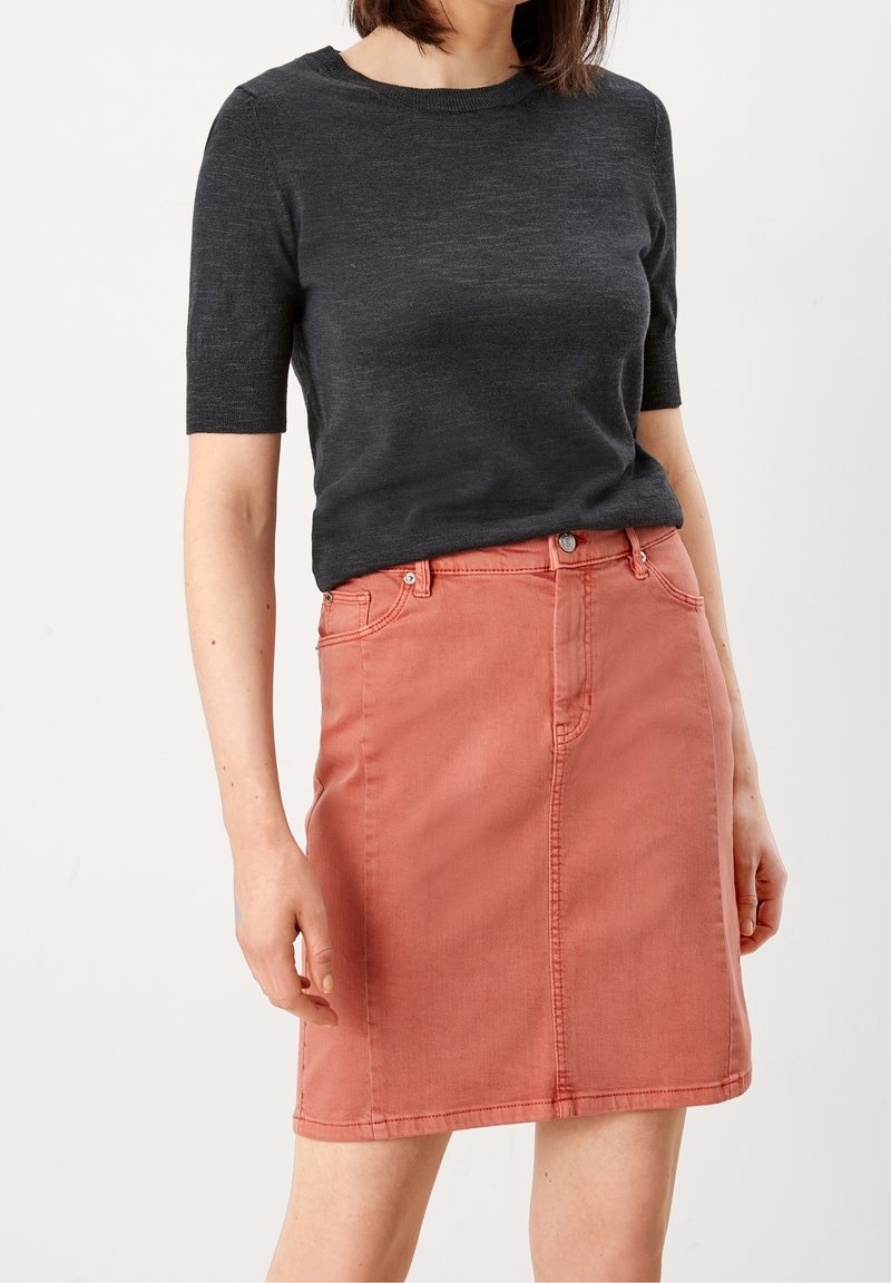 Femme portant un haut en maille gris foncé à manches courtes et une jupe trapèze en denim rose corail, debout devant un fond uni.