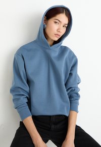 Modrá mikina s kapucí, volného střihu a dlouhými rukávy. Měkká tkanina bez vzorů. Model má na sobě černé džíny a sedí na bílém pozadí.