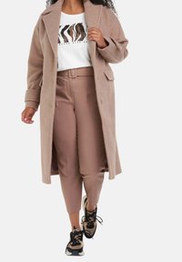 Lysbrun uldblanding frakke, hvid grafisk t-shirt, beige høj talje bukser og sorte sneakers med beige accenter og snørebånd.