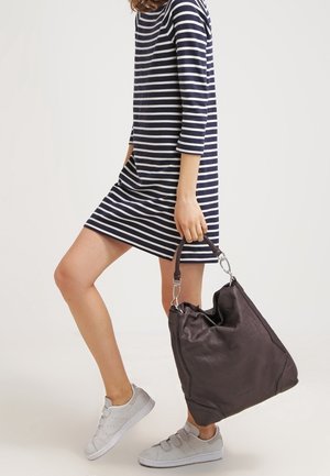 Kobieta w granatowo-białej sukience w paski, trzymająca dużą ciemnobrązową skórzaną torbę, ubrana w jasnoszare sneakersy.