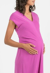 Äitiysmekko kirkkaassa vaaleanpunaisessa, jossa on V-kaula-aukko ja lyhyet hihat. Kangas on sileä ja valuu lempeästi pyöreän vatsan yli.