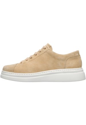 Beige suede sneaker with round toe, soft texture, six eyelets, and light beige laces. White rubber sole with subtle branding on the side.
