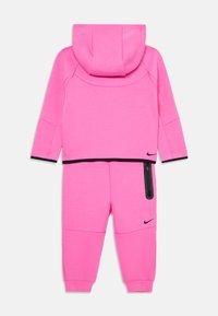 Różowy dres Nike z kapturem oraz spodniami z elastycznym pasem. Zawiera czarne akcenty, kieszeń na zamek błyskawiczny oraz miękką teksturę materiału.