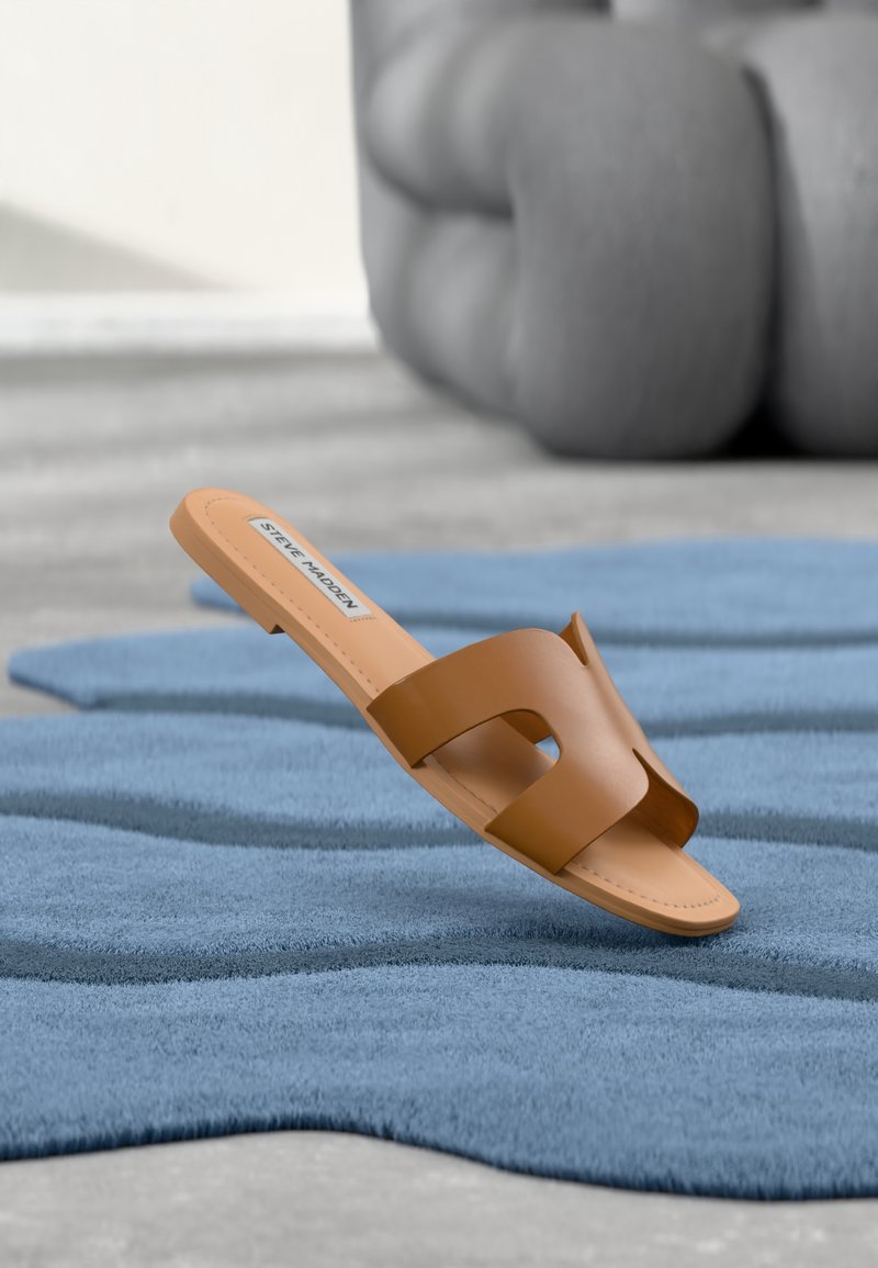 Sandale mule Steve Madden couleur tan à bout ouvert reposant sur un tapis bleu texturé avec un grand objet gris flou en arrière-plan.