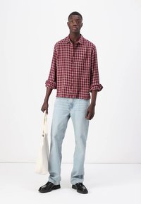 Rotschwarz-weiß karierte Hemd mit hochgerollten Ärmeln, kombiniert mit hellblauen Jeans und schwarzen Schuhen, dabei eine cremefarbene Stofftasche.