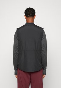 Schwarze ärmellose Weste aus leichtem Material, mit einem Kragen und glatter Textur, über einem dunklen Langarmshirt und burgunderfarbenen Hosen getragen.