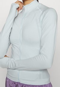 Světle modrá zipová bunda vyrobená z hladkého materiálu s vysokým límcem a dlouhými rukávy, obsahující jemné texturované panely a přiléhavý střih.