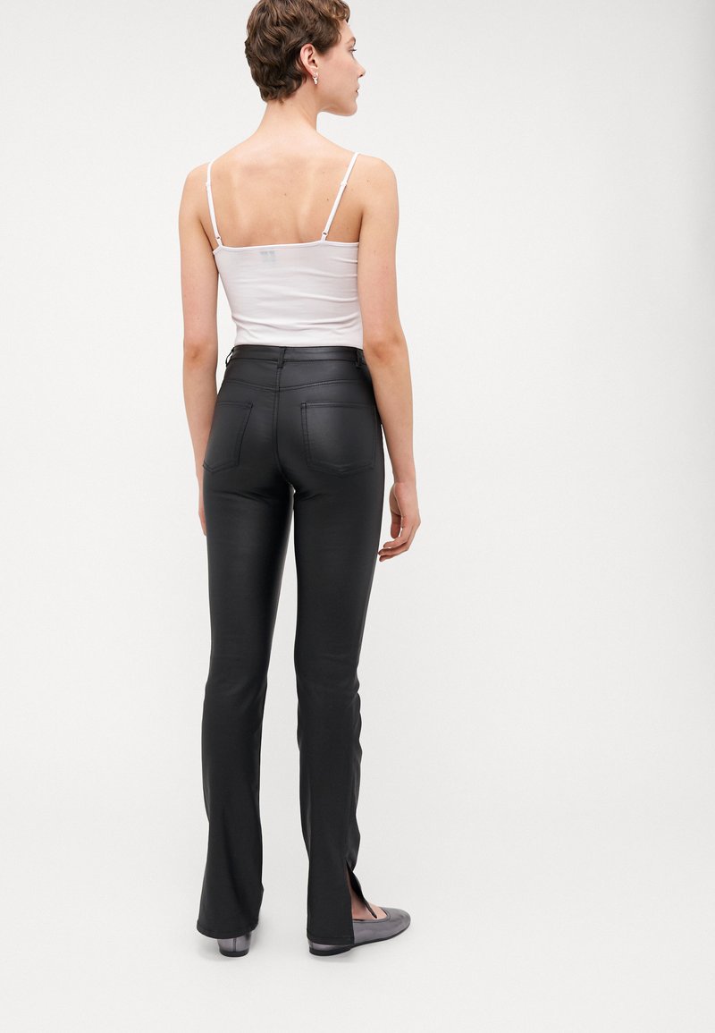 Schwarze, aus Kunstleder gefertigte, weit geschnittene Hose mit seitlichen Schlitzen, kombiniert mit einem weißen ärmellosen Oberteil. Einfaches Design und zwei Gesäßtaschen.