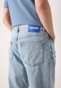 Jeans de mezclilla azul claro con un corte relajado, que presentan una etiqueta azul "HUGO" y un sutil detalle desgastado en los bolsillos traseros.