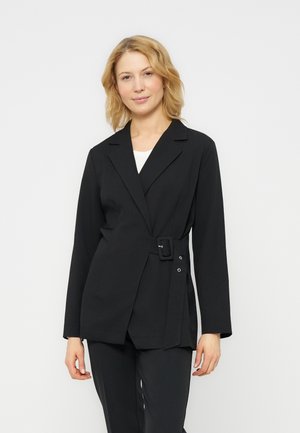 Femme aux cheveux blonds portant un blazer noir ceinturé sur un haut blanc et un pantalon noir, debout devant un fond blanc uni.