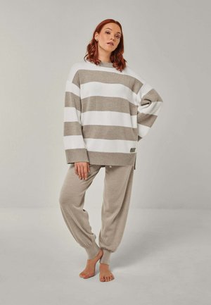 Woman standing barefoot wearing a beige and white wide-striped sweater and beige cuffed sweatpants against a plain background.