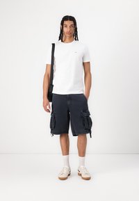 Witte katoenen t-shirt gecombineerd met donkere cargo shorts met zijzakken. Model draagt witte sokken en sneakers, en houdt een zwarte schoudertas vast.