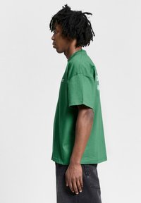 Camiseta verde de manga corta de algodón, con un escote redondo y un corte recto. Presenta el texto del logo en el lado frontal izquierdo y en la parte trasera derecha.