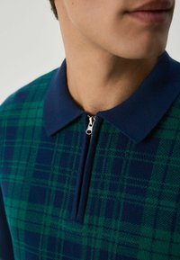 Nahaufnahme einer Person, die einen navyblauen und grünen karierter Reißverschluss-Pullover mit umgeschlagenem Kragen trägt.