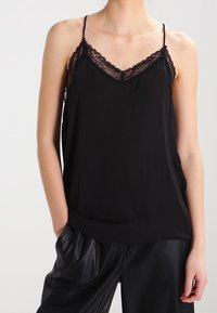 Svart linne med spetsdetaljer vid uttycket, tunna justerbara axelband och en slät tygfinish, matchat med löst sittande svarta läderbyxor.