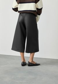 Person, die schwarze, weit geschnittene, verkürzte Hose, schwarze spitze Ballerinas und einen braun, creme- und schwarzgestreiften Pullover trägt und auf einem grauen Boden steht.