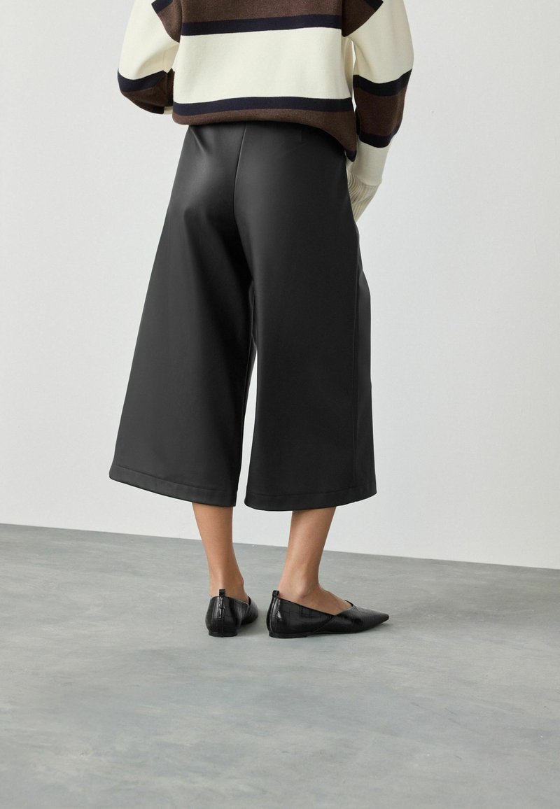 Person, die schwarze, weit geschnittene, verkürzte Hose, schwarze spitze Ballerinas und einen braun, creme- und schwarzgestreiften Pullover trägt und auf einem grauen Boden steht.