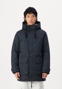 Parka de invierno en color azul marino con capucha, dos grandes bolsillos frontales, cierres de botones a presión y cremallera, acentos en negro y una superficie texturizada.