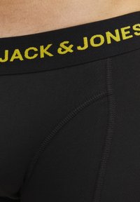 Schwarze Boxer-Shorts mit einem gelben "JACK & JONES"-Logo auf dem elastischen Bund. Glatte Stoffoberfläche und kontrastierende Nähte.