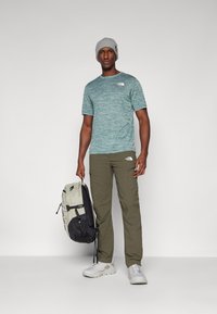 Roheline täpiline t-särk, oliivivärvi cargo-püksid, hall beanie ja hallid kingad. Hoides musta ja heleda halli seljakotti, millel on mitu taskut.