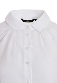 Biała bluzka z kołnierzykiem, z siedmioma guzikami z przodu, z falbaniastym detalem i gładką, błyszczącą teksturą materiału.