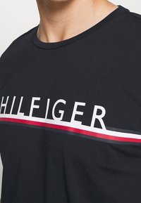 Detailní pohled na osobu oblečenou v černém tričku s bílými písmeny "HILFIGER" a červenými, bílými a modrými vodorovnými pruhy přes hrudník.