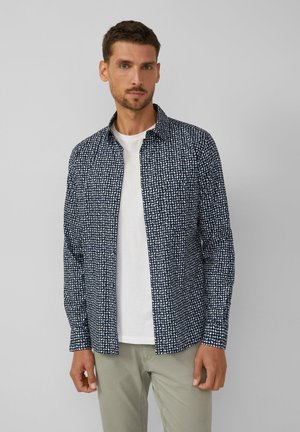 Man in een open marineblauw overhemd met een wit stippenpatroon over een wit T-shirt en lichte grijze broek, staand tegen een effen grijze achtergrond.