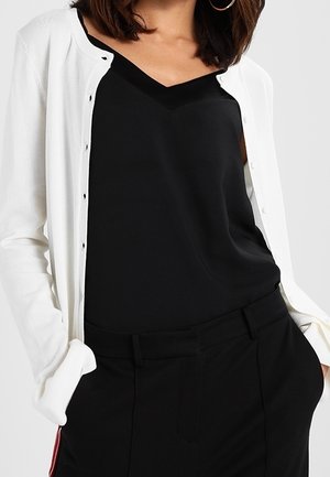 Femme portant un cardigan blanc boutonné par-dessus un haut noir à col en V et un pantalon noir, les mains dans les poches.