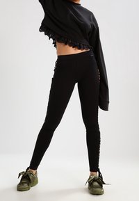 Felpa nera corta con bordi in pizzo e leggins neri con aperture laterali; abbinati a scarpe da ginnastica verde oliva con fiocchi.