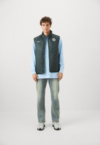 Mørkegrøn quiltet puffer vest med Nike-logo og Chelsea FC-emblemet, båret over en lys blå langærmet skjorte, parret med slidte jeans og sneakers.