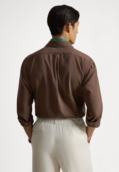 Καφέ πουκάμισο με μακριά μανίκια, διπλωμένο γιακά και κουμπωτά μανσέτες, με πτυχώσεις στο πίσω μέρος. Συνδυασμένο με ανοιχτόχρωμο, υφασμάτινο παντελόνι.